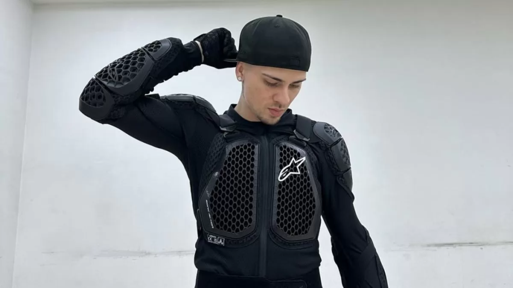 El cantante urbano chileno Easykid posa mirando hacia abajo, vistiendo una gorra negra y una armadura de protección de motociclista.