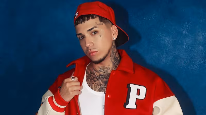 
Young Cister posa frente a un fondo azul vistiendo una chaqueta roja y blanca, una camiseta blanca y una gorra roja hacia atrás. El artista mira directamente a la cámara mientras muestra sus tatuajes en el cuello y el rostro.
