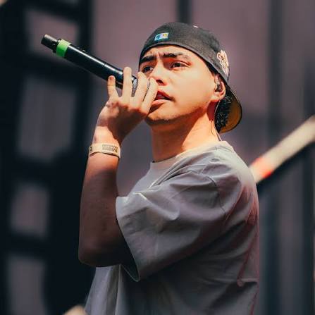 Fotografía de Pepe Fly actuando en el escenario. Lleva una camiseta clara, una gorra negra al revés y está cantando con un micrófono. El fondo está borroso.