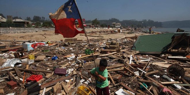 Niño pequeño cargando una bandera nacional en medio de escombros a la orilla del mar.