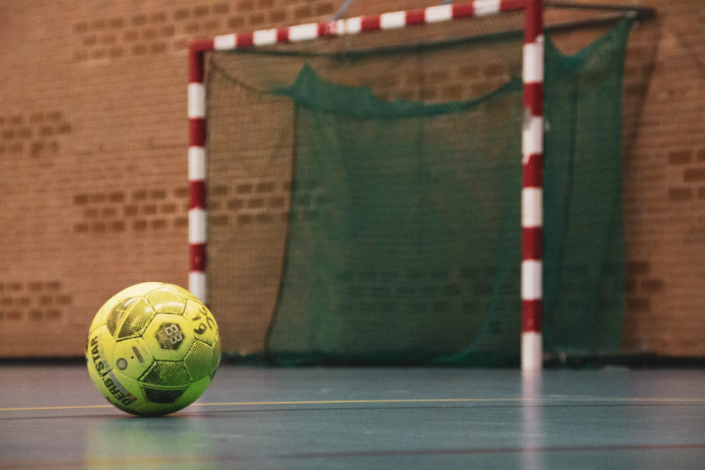 En esta imagen se puede visualizar un balón de Futsal, atrás de este se encuentra un arco de Fustal.