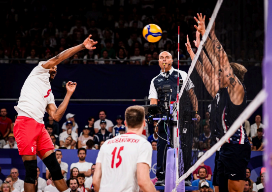 En esta imagen se ve un juego de voleibol, centrándose en el saque de partida del equipo de blanco con rojo hacia el equipo defensor del color azul. Todo esto presenciado por un arbitro.
